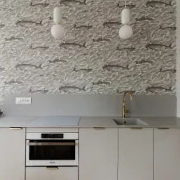 Kuchnia w odcieniach szarości z tapetą w wieloryby, złota bateria i parkiet w jodełkę, oświetlona dwoma wiszącymi lampami z kulistymi kloszami.