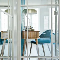 Nowoczesny gabinet za szklanymi drzwiami: drewniane biurko z lampką i książkami, welurowe krzesło w odcieniach błękitu, dywan w geometryczny wzór, jasne wnętrze z oknami.