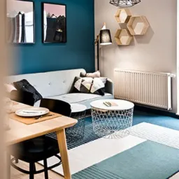 Salon w Gdyni z turkusową ścianą, szarą sofą, geometrycznym dywanem i lampą w kształcie klatki, uzupełniony drewnianymi półkami w kształcie plastrów miodu i okrągłym stolikiem kawowym z metalową...