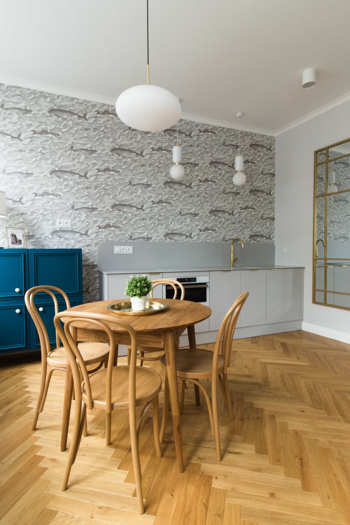 Jadalnia z okrągłym, drewnianym stołem i krzesłami w stylu retro, na blacie złota taca z małą rośliną doniczkową, w tle ściana z tapetą w motywie ryb, błękitny kredens i złote lustro.