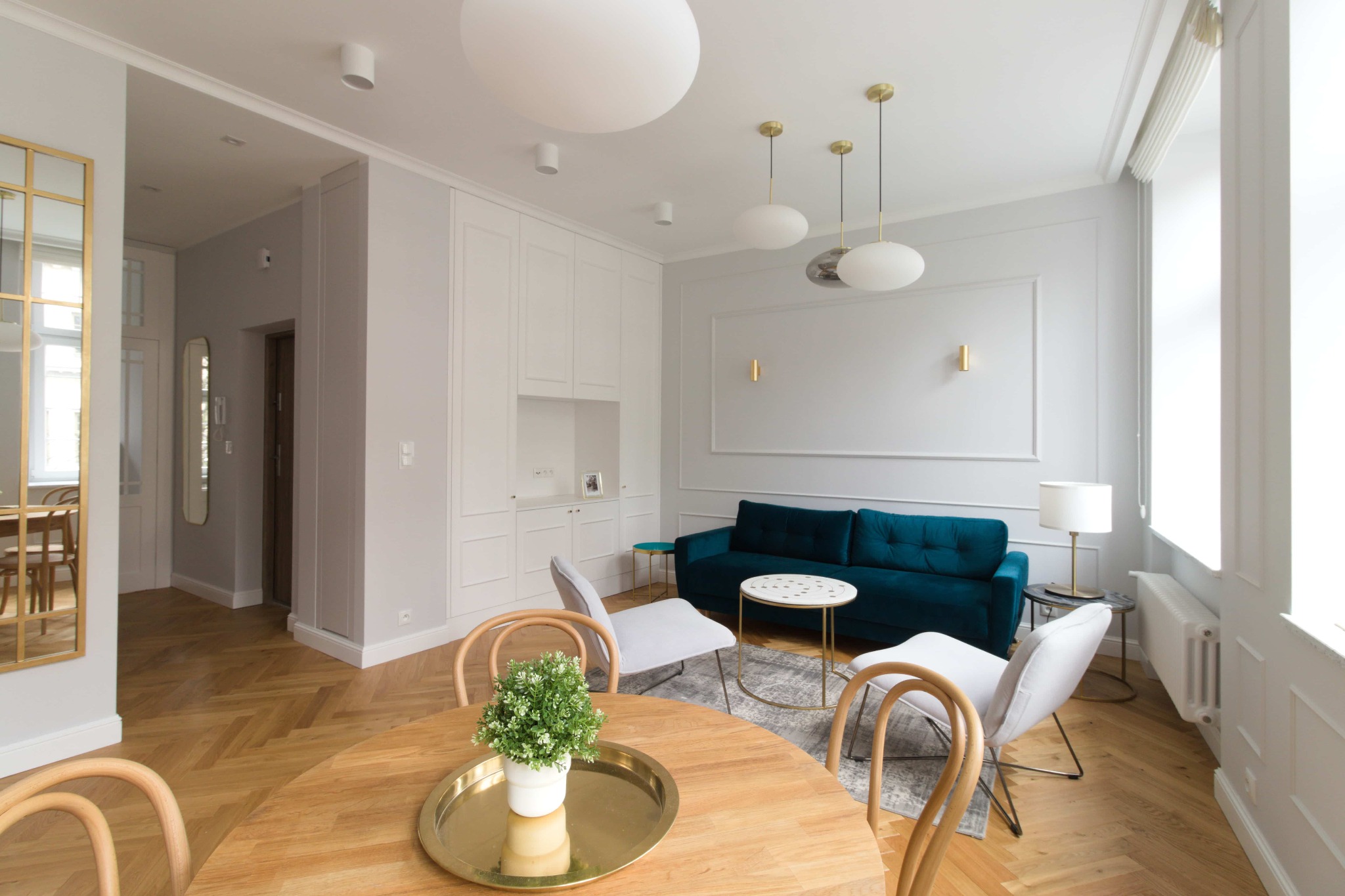 Jasne wnętrze z drewnianą podłogą w jodełkę, okrągły stół z rośliną w doniczce na złotej tacy, turkusowa sofa i białe krzesła, oświetlenie sufitowe w formie kul, ściany w odcieniach bieli...