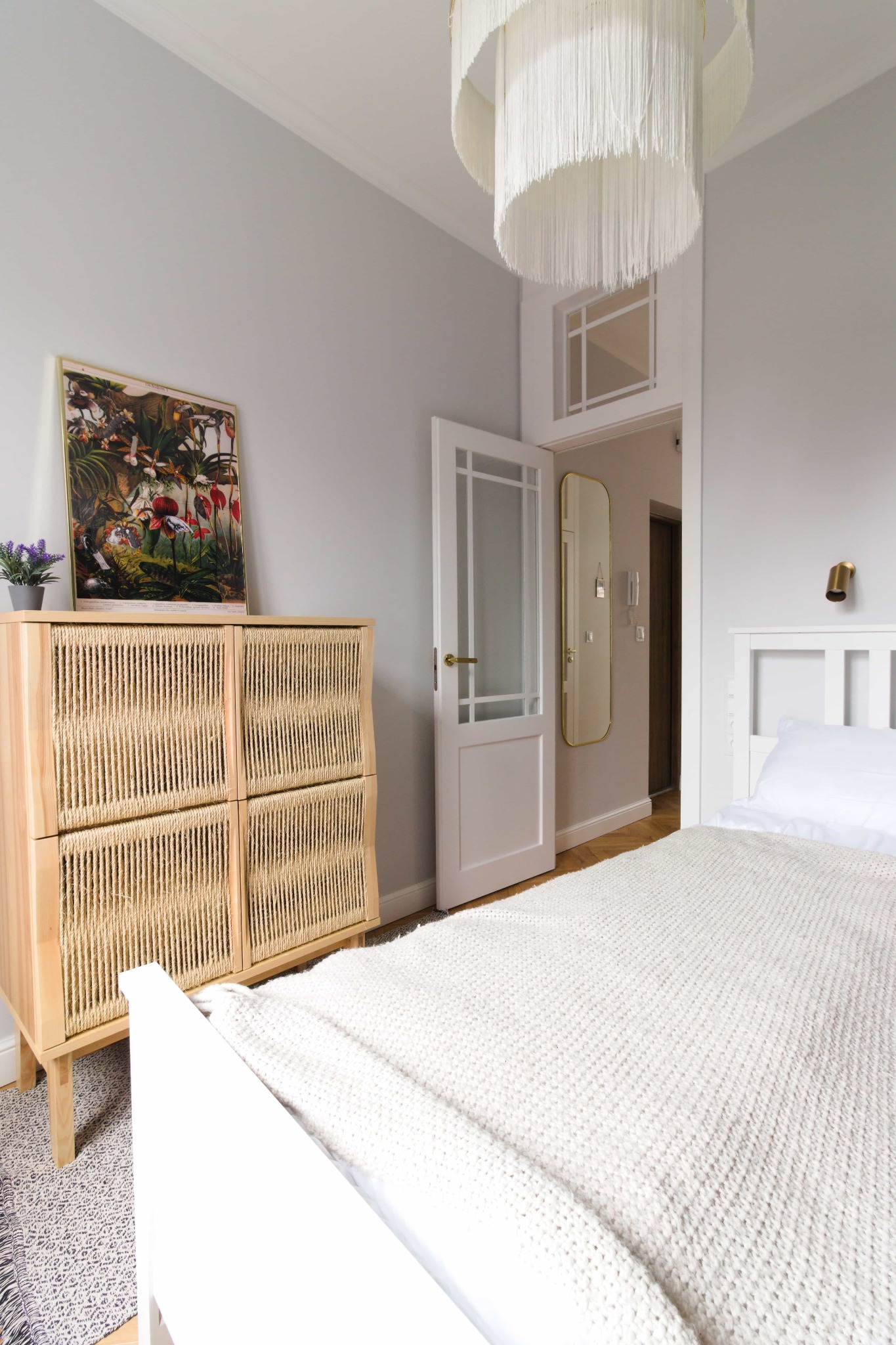 Jasna sypialnia z białym łóżkiem, komodą z plecionki rattanowej, wiszącą lampą z frędzlami i otwartymi białymi drzwiami do przedpokoju z lustrem.