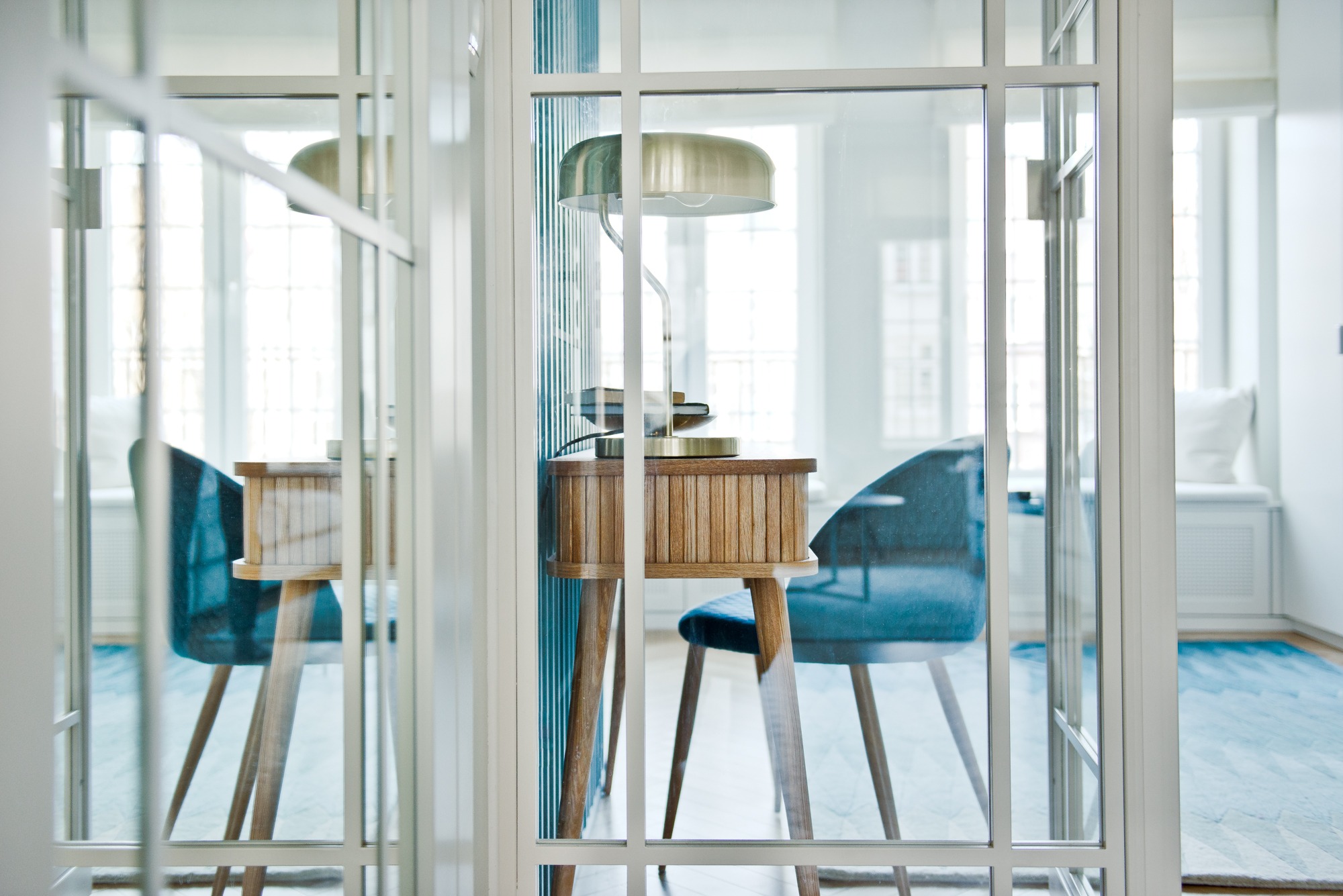 Nowoczesny gabinet za szklanymi drzwiami: drewniane biurko z lampką i książkami, welurowe krzesło w odcieniach błękitu, dywan w geometryczny wzór, jasne wnętrze z oknami.