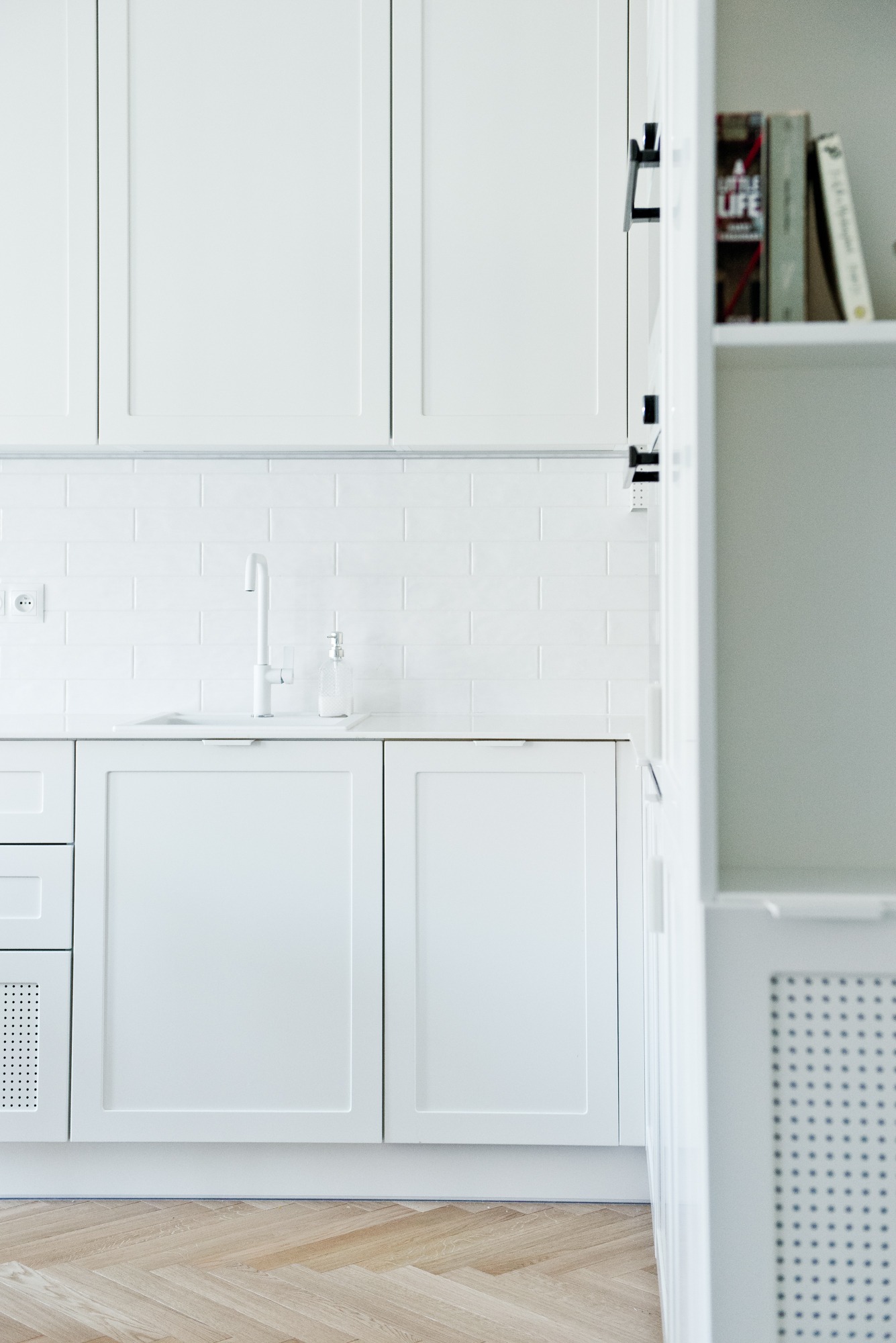 Minimalistyczna, biała kuchnia z drewnianą podłogą w jodełkę, bateria i dozownik na blacie, płytki metro na ścianie.