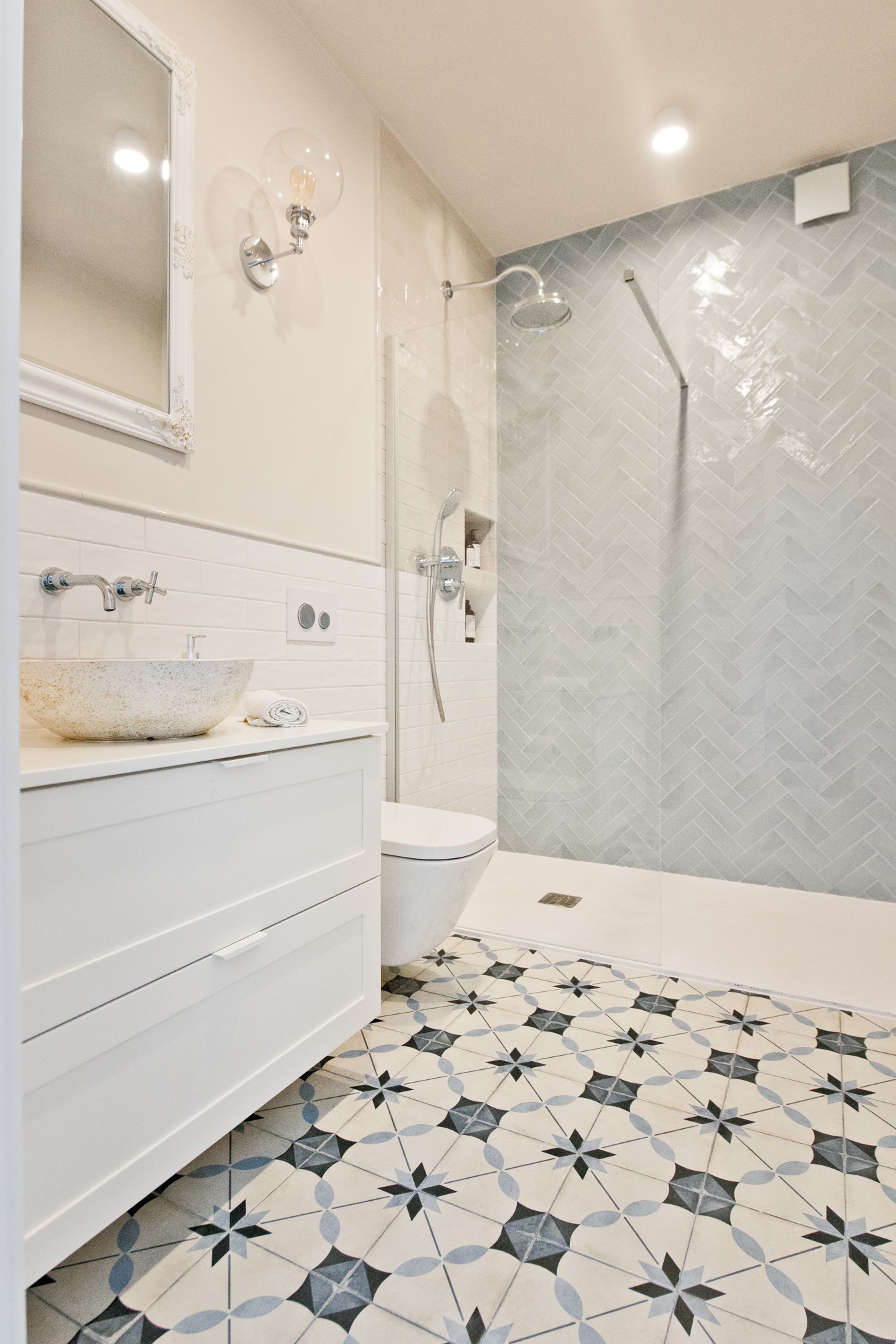 Jasna łazienka z wzorzystą podłogą, kamienną umywalką na białej szafce i prysznicem z szarymi płytkami ułożonymi w jodełkę.