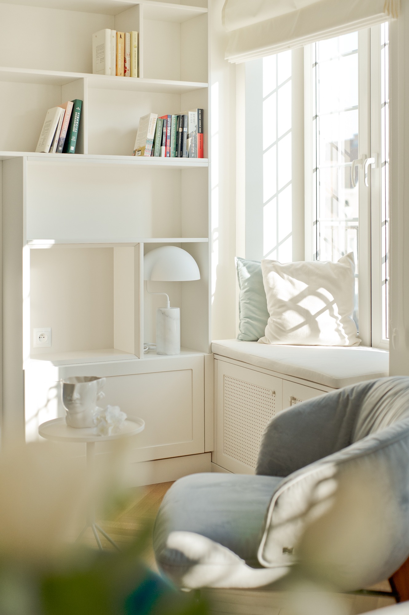 Jasne wnętrze z białymi regałami, siedziskiem pod oknem z poduszkami i szarym fotelem, oświetlone naturalnym światłem wpadającym przez okno.