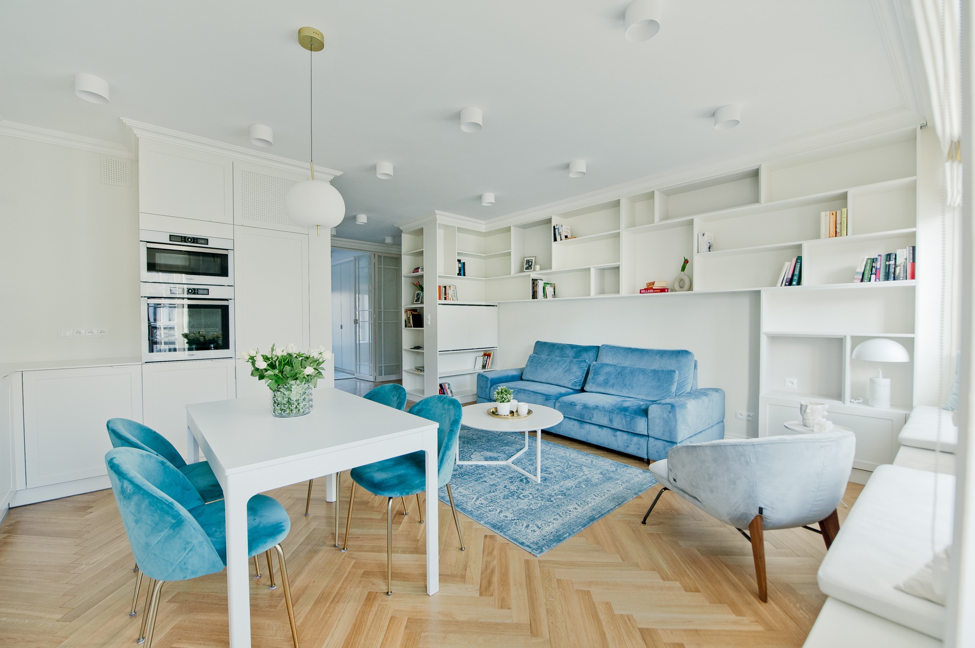 Jasne, przestronne wnętrze z parkietem w jodełkę, błękitną sofą i krzesłami z aksamitu, białą zabudową kuchenną z piekarnikiem i mikrofalówką, oraz regałem z książkami w tle.