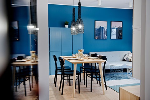 Niebieski salon z jadalnią, drewniany stół z czarnymi krzesłami, rattanowe lampy wiszące, szara sofa z poduszkami, obrazy górskie na ścianie, widok przez drzwi.