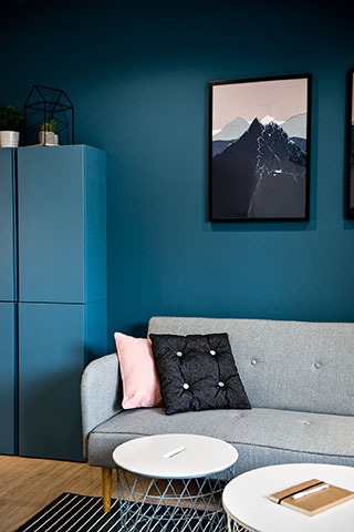 Nowoczesny salon z turkusowymi ścianami, szarą sofą z różową i czarną poduszką, dwoma okrągłymi stolikami kawowymi i abstrakcyjnymi obrazami górskimi na ścianie.