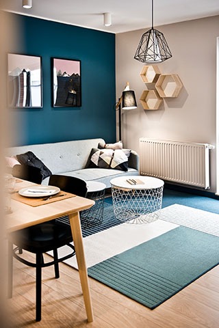 Salon w Gdyni z turkusową ścianą, szarą sofą, geometrycznym dywanem i lampą w kształcie klatki, uzupełniony drewnianymi półkami w kształcie plastrów miodu i okrągłym stolikiem kawowym z metalową...