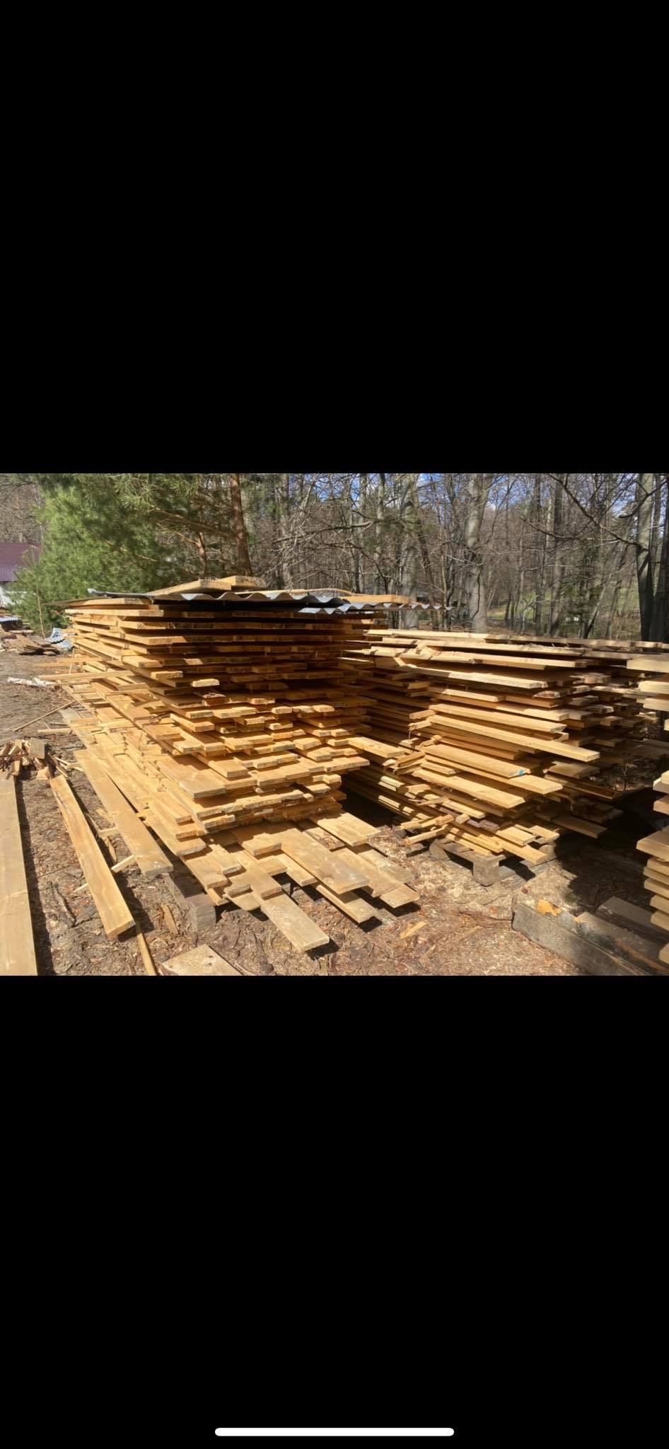 Stosy świeżo przetartego drewna ułożone na zewnątrz, zabezpieczone blachą falistą, z widocznym lasem w tle.