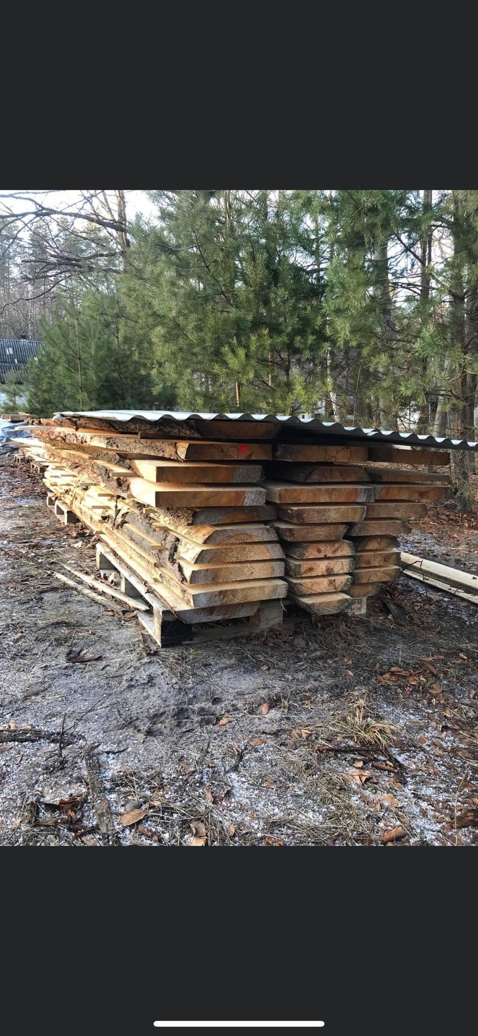 Stos desek z surowego drewna ułożonych na palecie, przykrytych blachą falistą, na tle drzew i ziemi pokrytej resztkami śniegu.