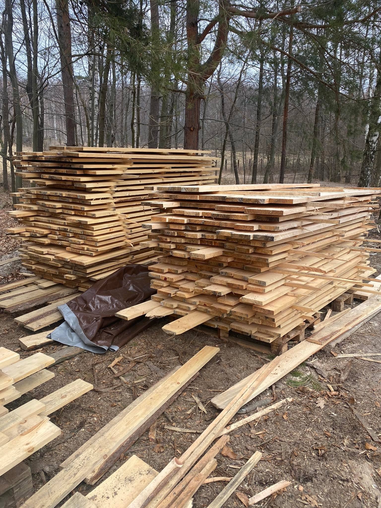 Dwa stosy świeżo przetartej tarcicy na paletach, częściowo przykryte plandeką, na ziemi z liśćmi i gałązkami, w tle las.