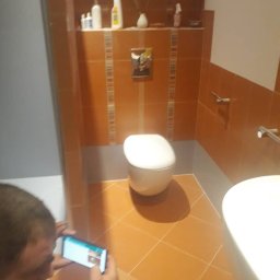 Benefits Aslan Bagaev - Wykończona łazienka z pomarańczowymi płytkami, białą toaletą i umywalką, fotografowana smartfonem przez osobę z boku.