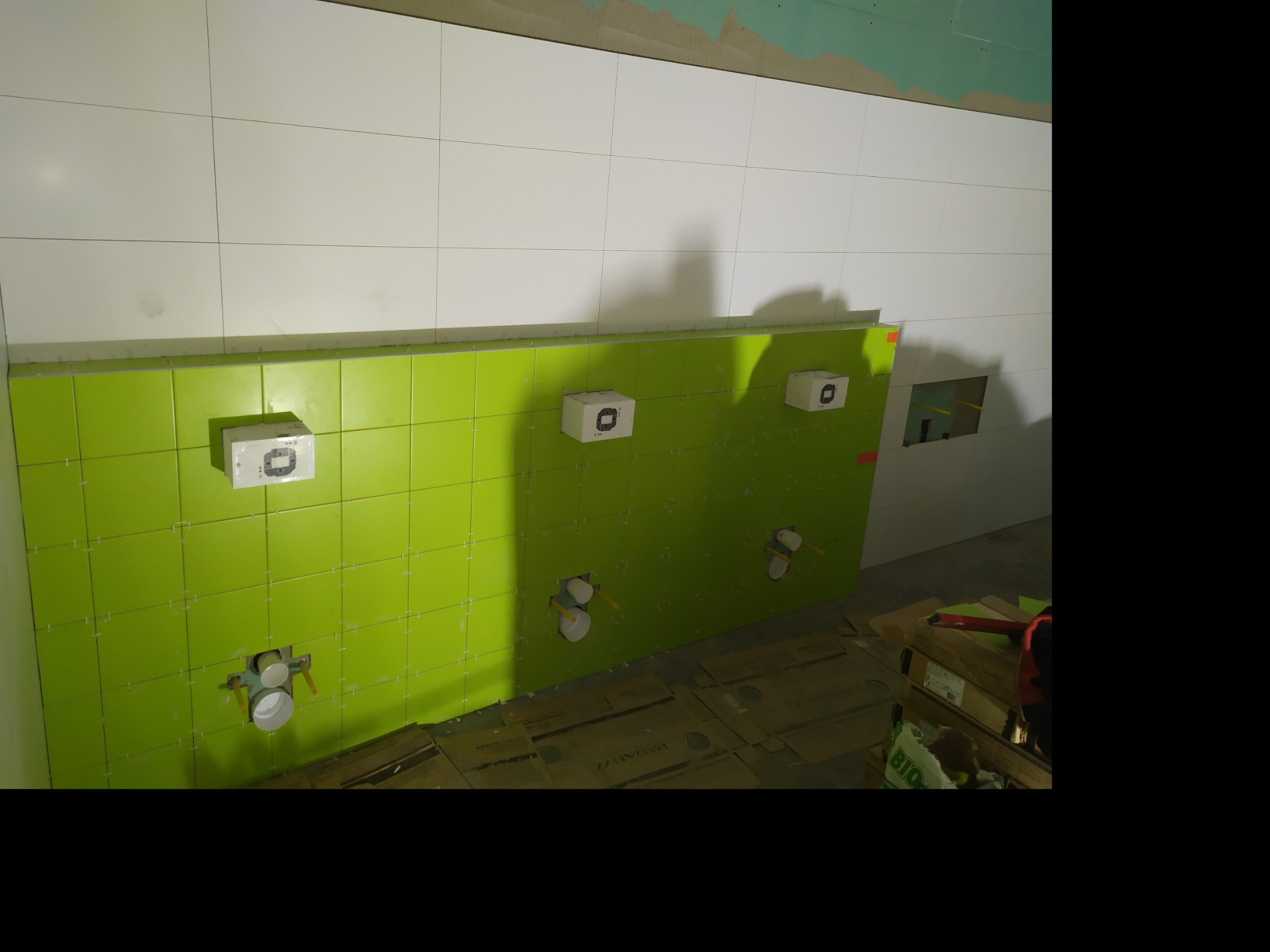 Ściana w trakcie kładzenia płytek: białe płytki na górze, zielone na dole, widoczne puszki instalacyjne i rury doprowadzające wodę, zabezpieczone kartonem podłoże.