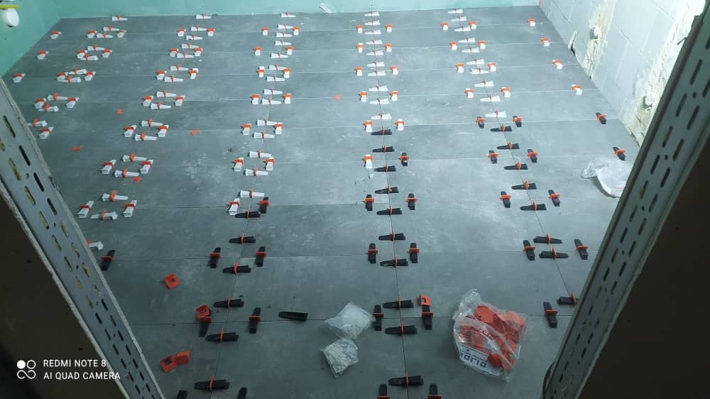 Układanie szarych płytek podłogowych z użyciem systemu poziomowania płytek (klipsy i kliny) w trakcie remontu.