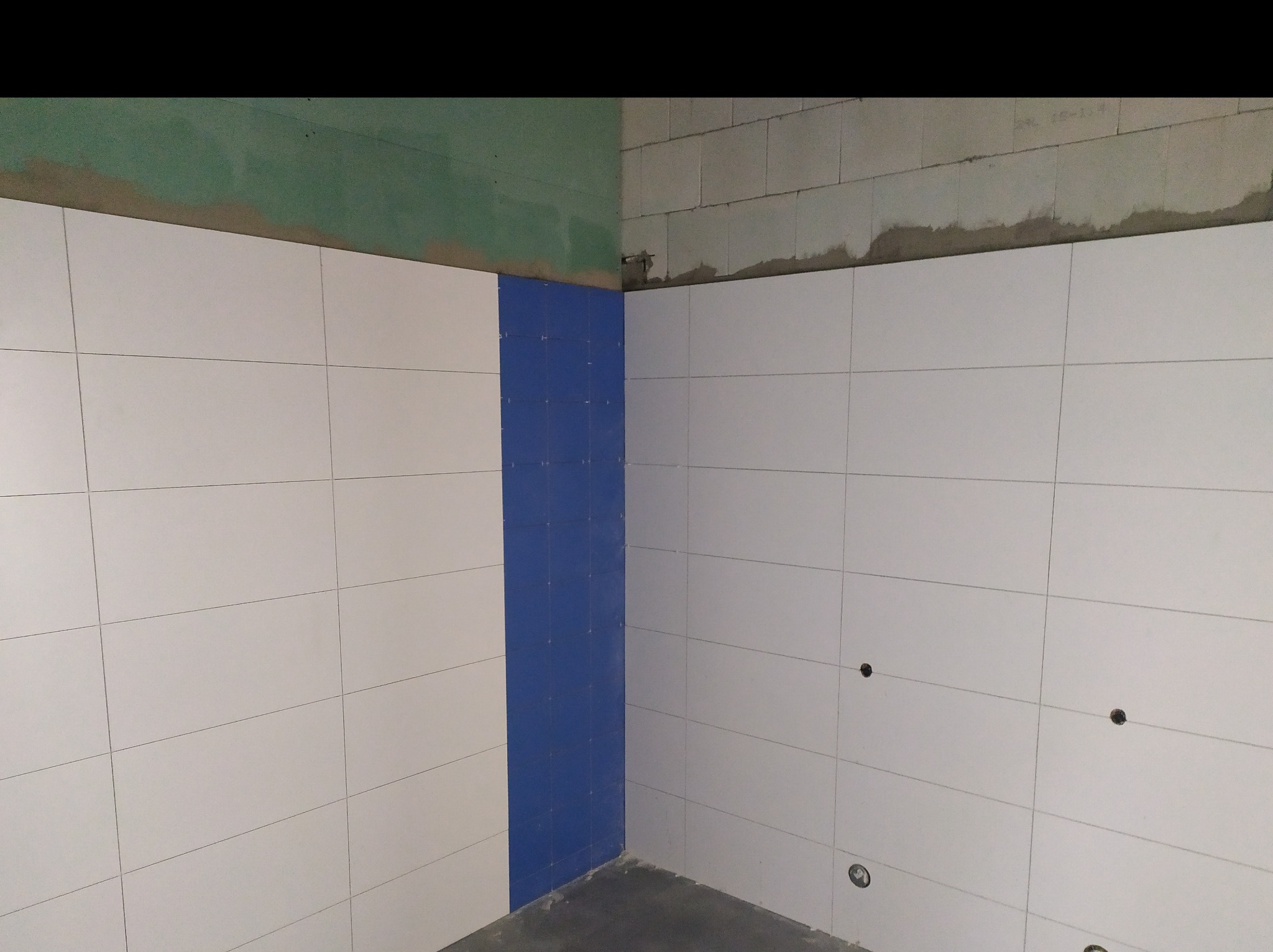 Układanie białych płytek na ścianie z pionowym pasem niebieskich płytek; widoczne otwory na instalacje, surowy stan ścian powyżej płytek.