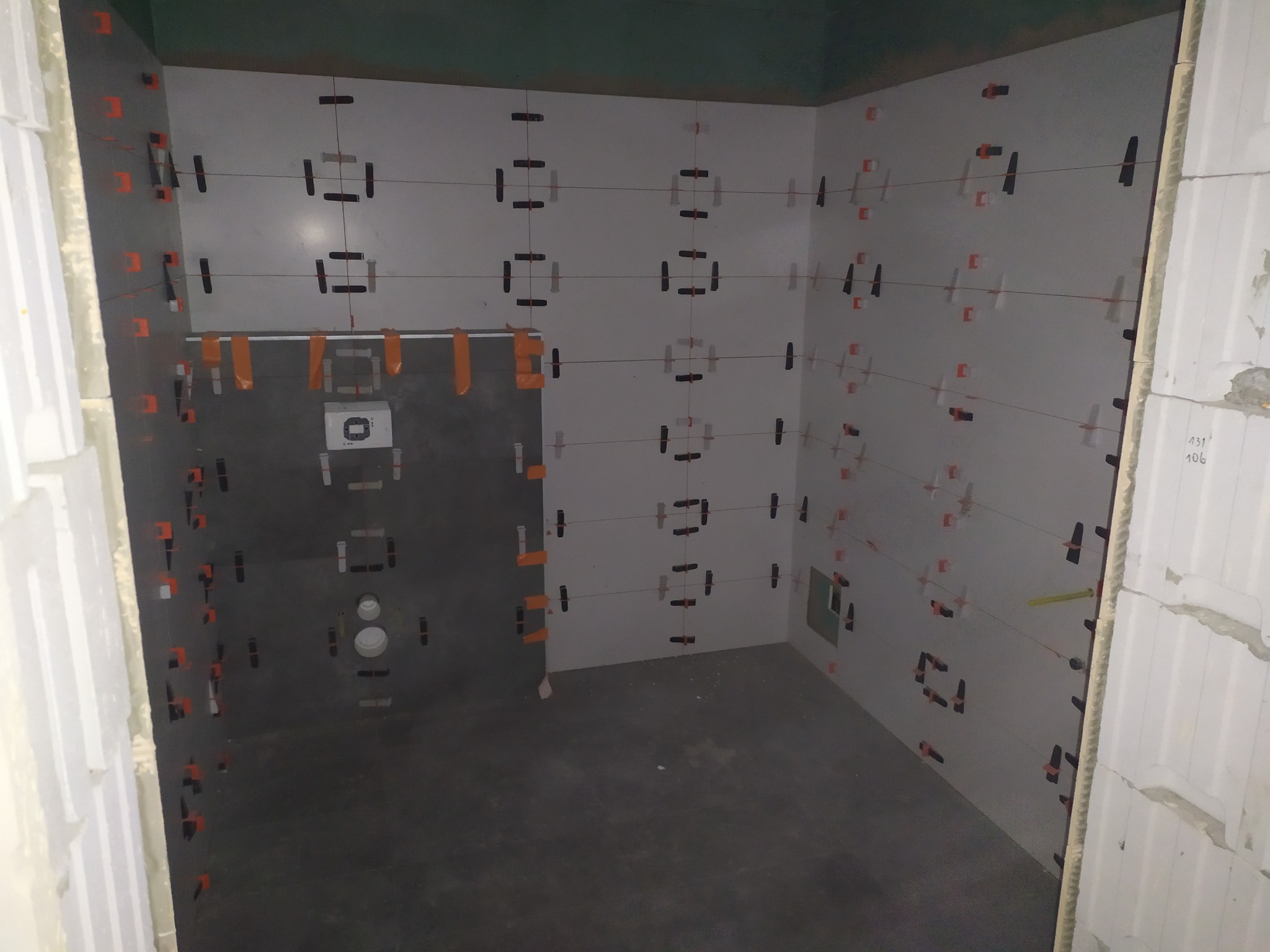 Wnętrze łazienki w trakcie układania jasnych płytek, widoczne krzyżyki dystansowe i poziomowanie sznurkiem, ciemna podłoga, ściany częściowo przygotowane do położenia płytek.