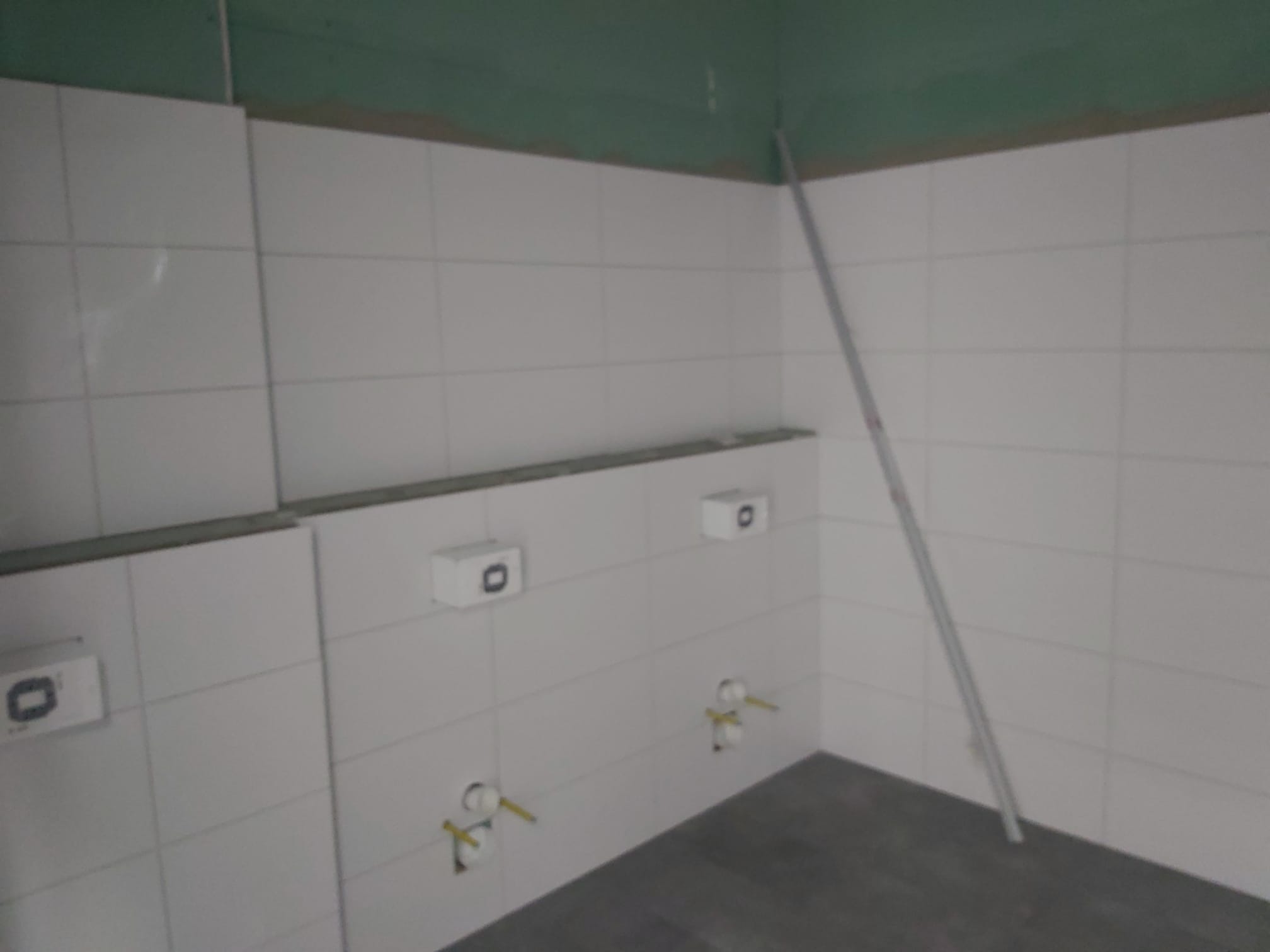 Białe płytki ceramiczne ułożone na ścianie w trakcie remontu łazienki, widoczne wystające rury instalacji wodnej i poziomica oparta o ścianę.