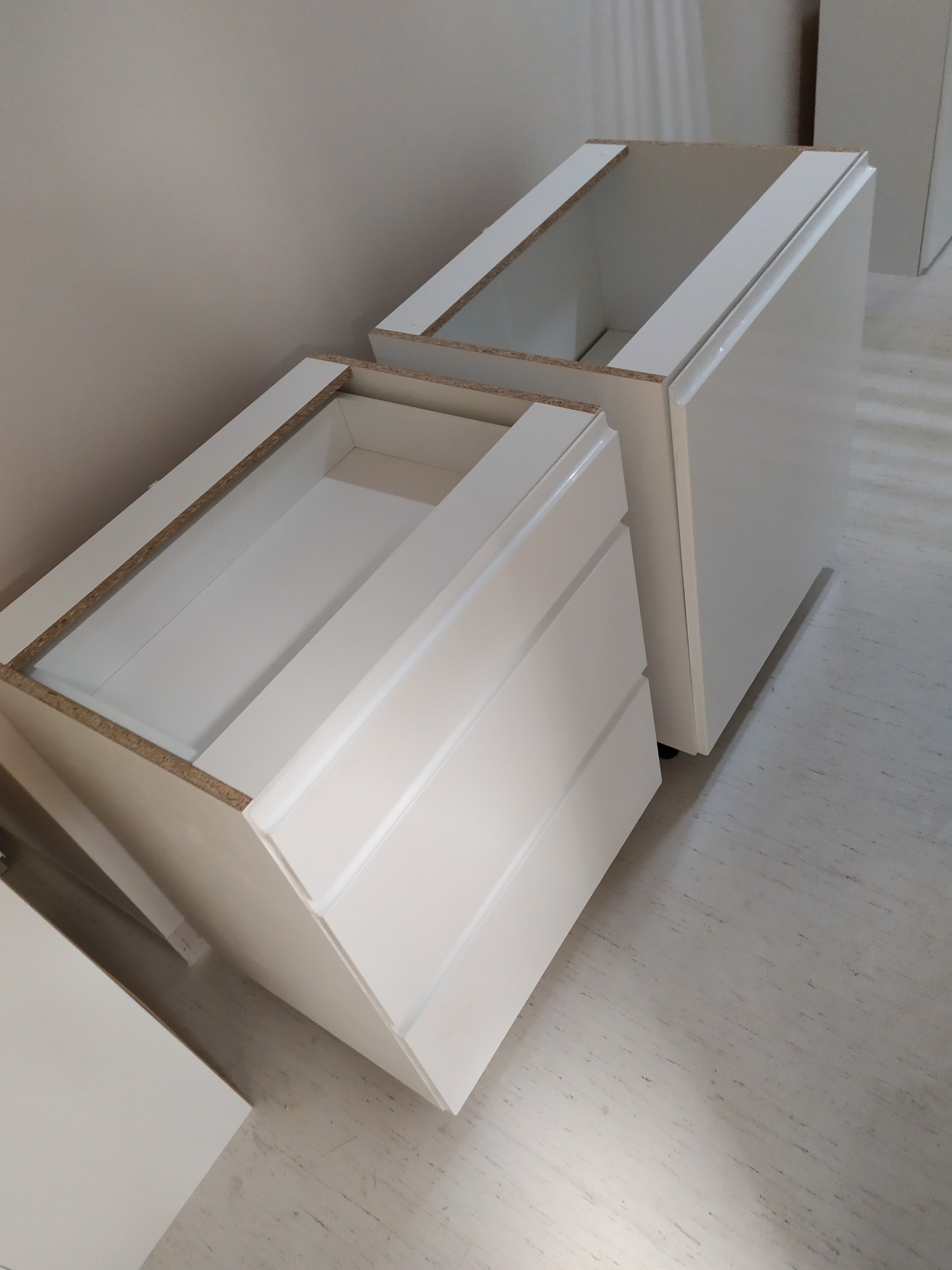 Dwa białe korpusy szafek kuchennych bez frontów, z widocznymi szufladami w jednym z nich, ustawione na podłodze w trakcie montażu.