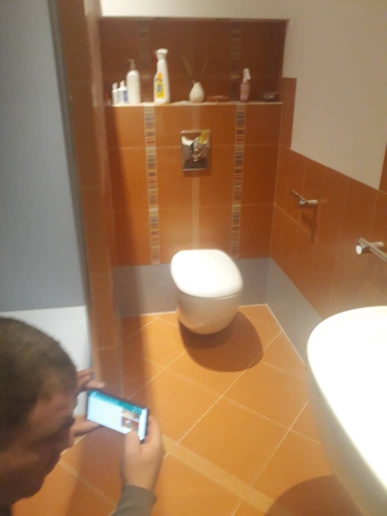 Wykończona łazienka z pomarańczowymi płytkami, białą toaletą i umywalką, fotografowana smartfonem przez osobę z boku.