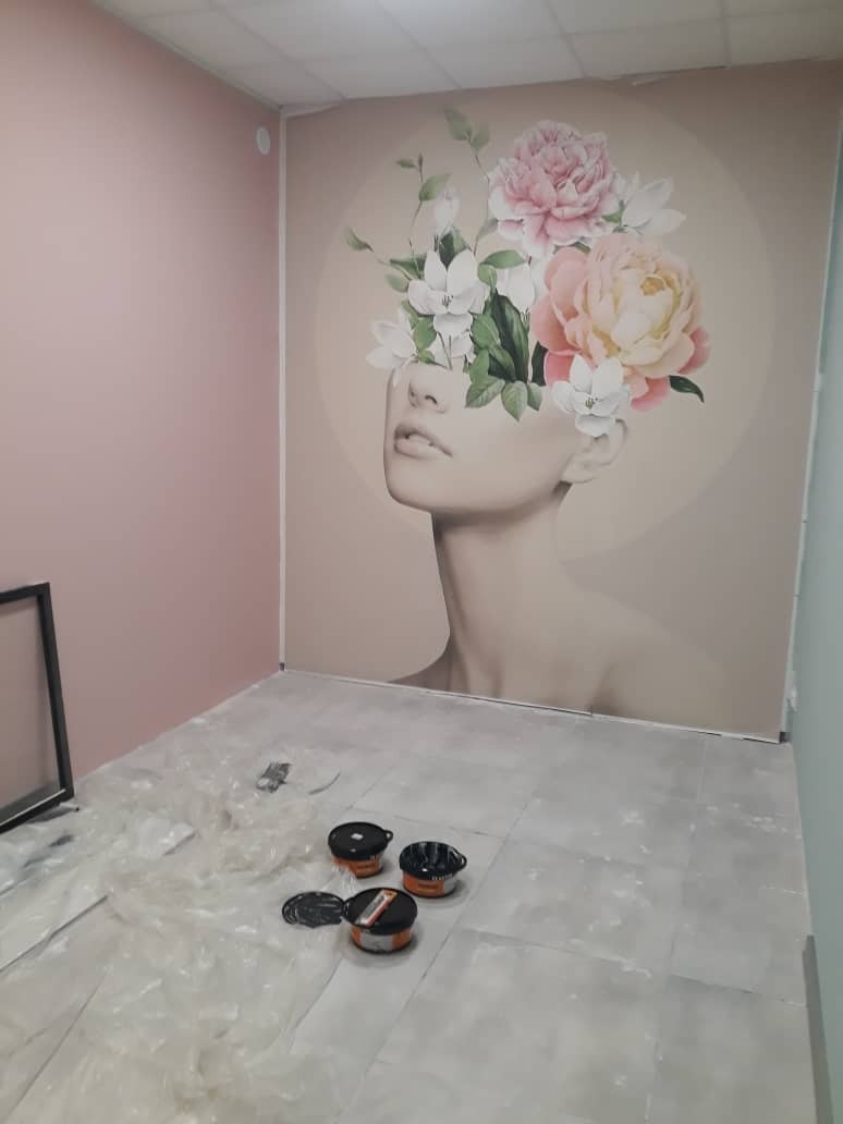 Świeżo pomalowane ściany w pokoju z dekoracyjnym muralem przedstawiającym portret kobiety z kwiatami zasłaniającymi oczy, widoczne puszki z farbą i folia ochronna na podłodze.