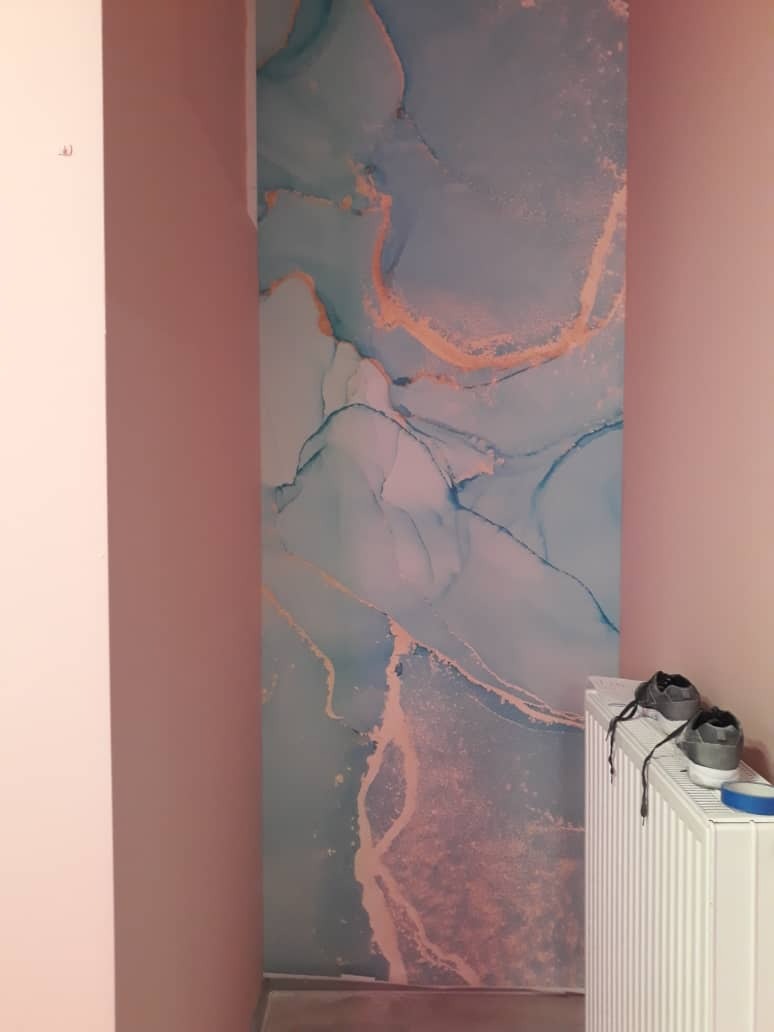 Ściana z tapetą w abstrakcyjny wzór akwareli w odcieniach błękitu i złota, fragment grzejnika z butami na górze, różowe ściany w tle.