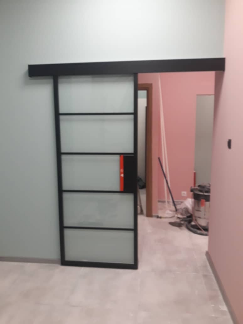 Zamontowane przesuwne drzwi szklane z czarną ramą, widoczne niedoskonałości wykończenia ścian i podłogi, odkurzacz przemysłowy w tle.