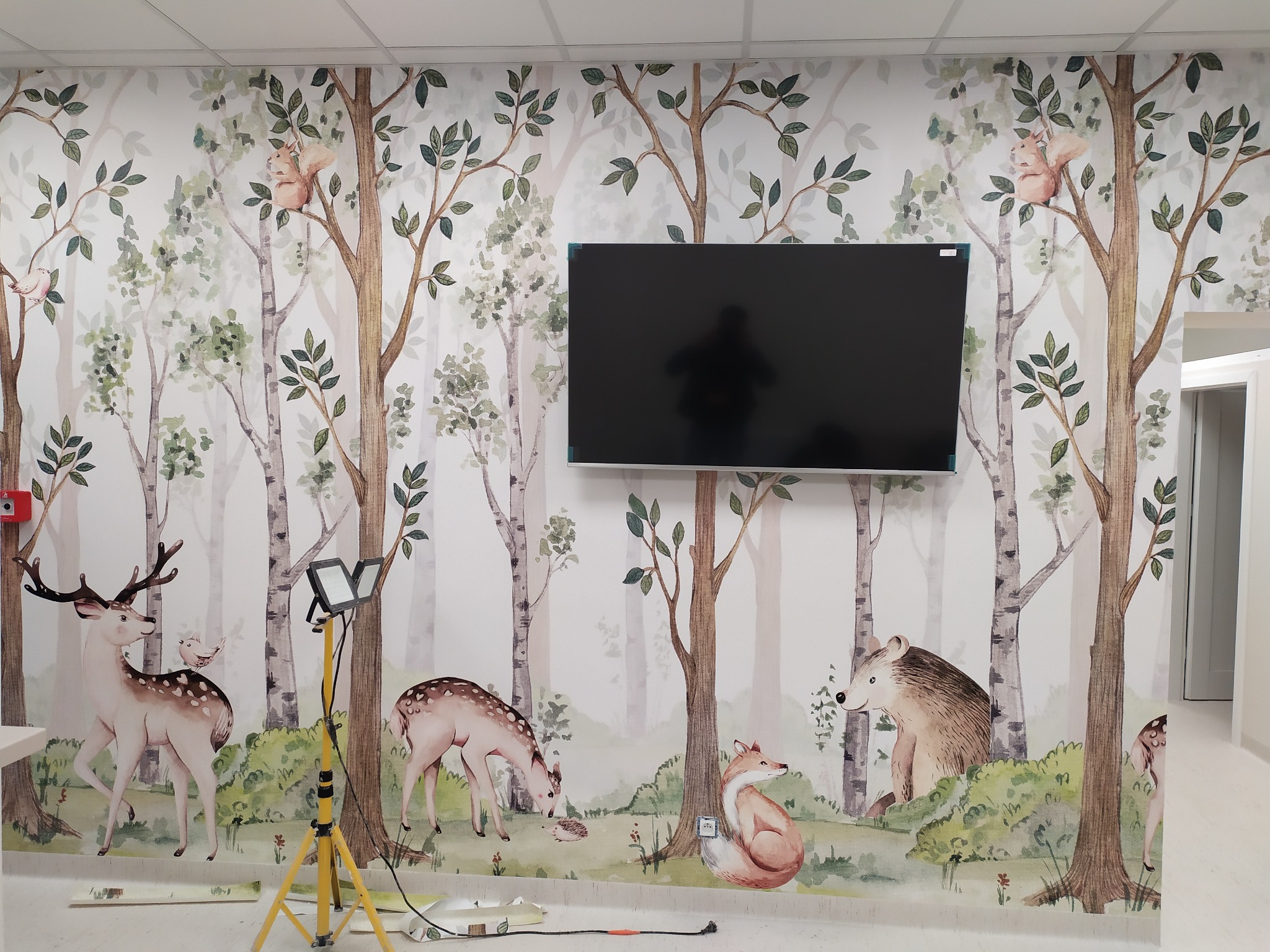 Ściana w pokoju dziecięcym pokryta fototapetą z motywem leśnym, przedstawiającą drzewa i zwierzęta, z zamontowanym telewizorem i lampą budowlaną na statywie.