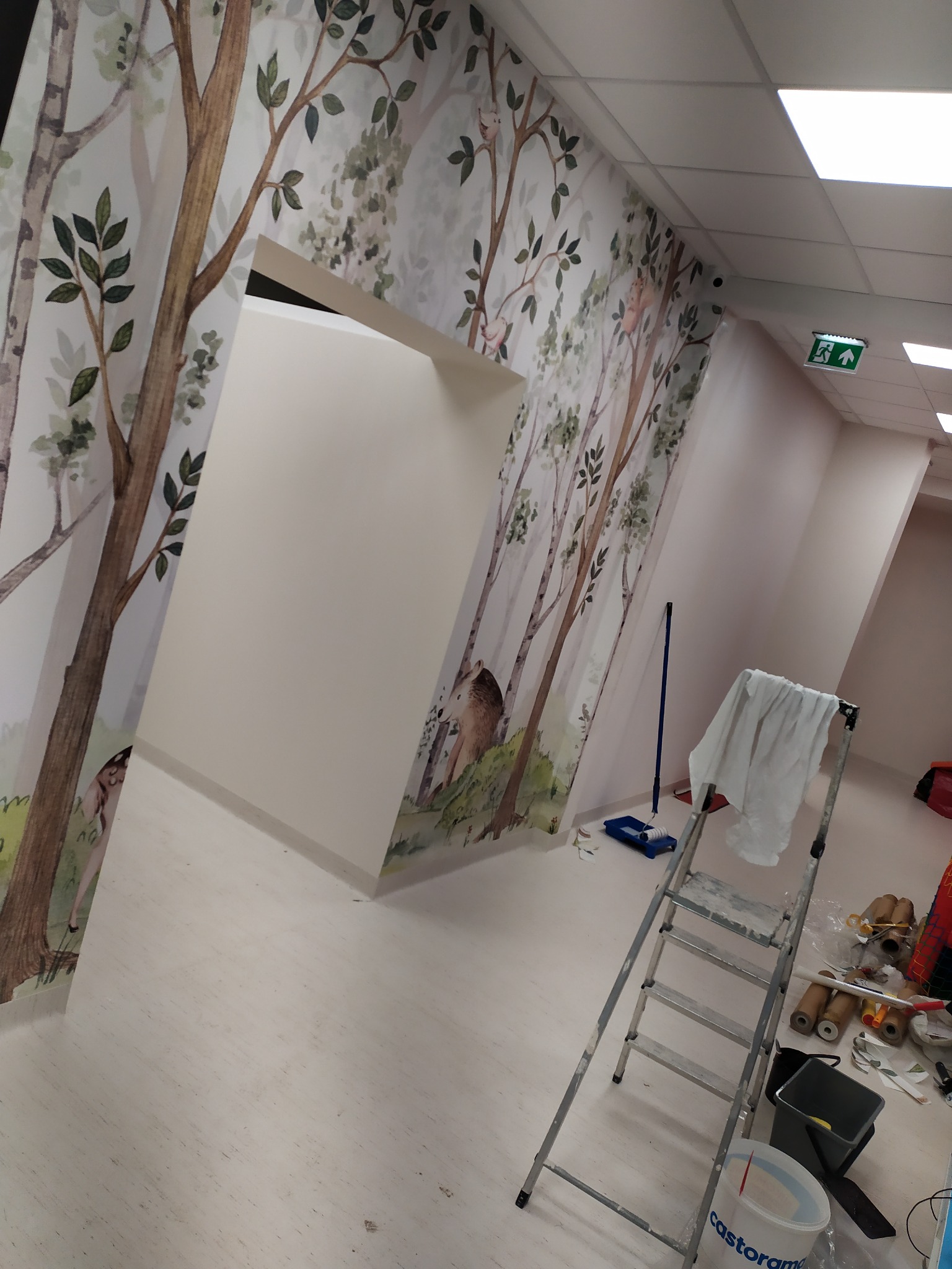 Pokój biurowy z fototapetą przedstawiającą las, drabina i narzędzia malarskie sugerują prace wykończeniowe.