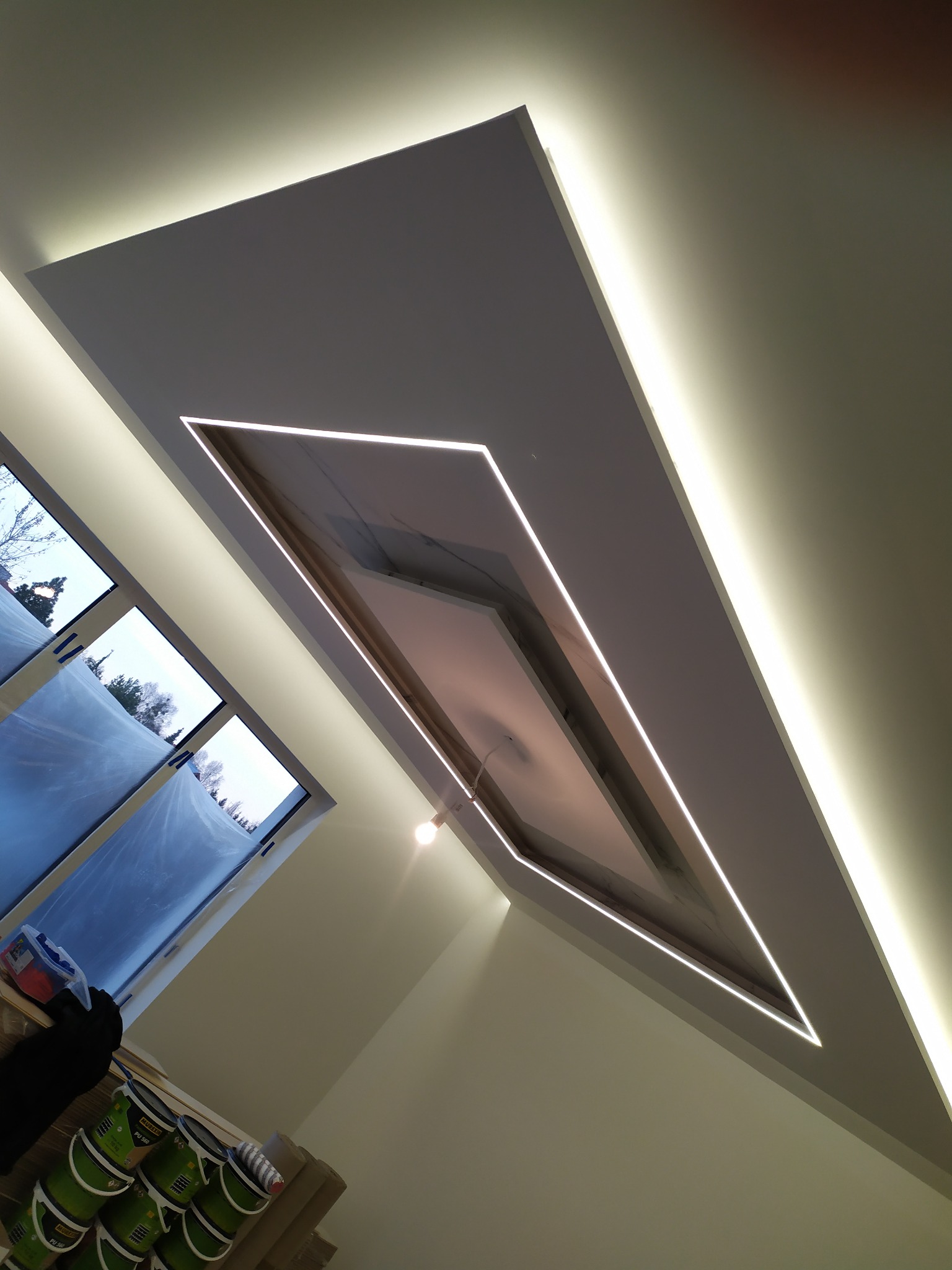 Nowoczesny sufit z podwieszaną konstrukcją i oświetleniem LED w kształcie rombu, widok z dołu, okna zakryte folią malarską w tle.