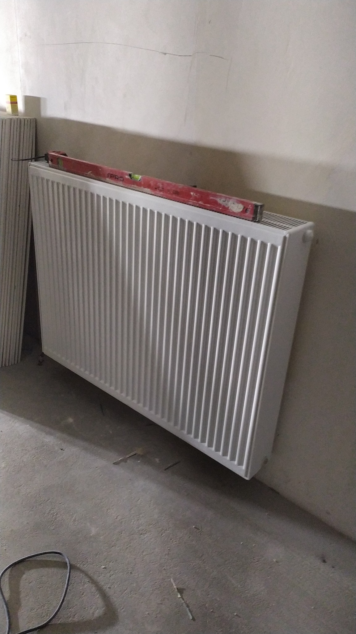Nowy, biały grzejnik panelowy zamontowany przy ścianie, poziomowany czerwoną poziomicą, widoczne ślady po gładzi na ścianie i niedokończona podłoga.