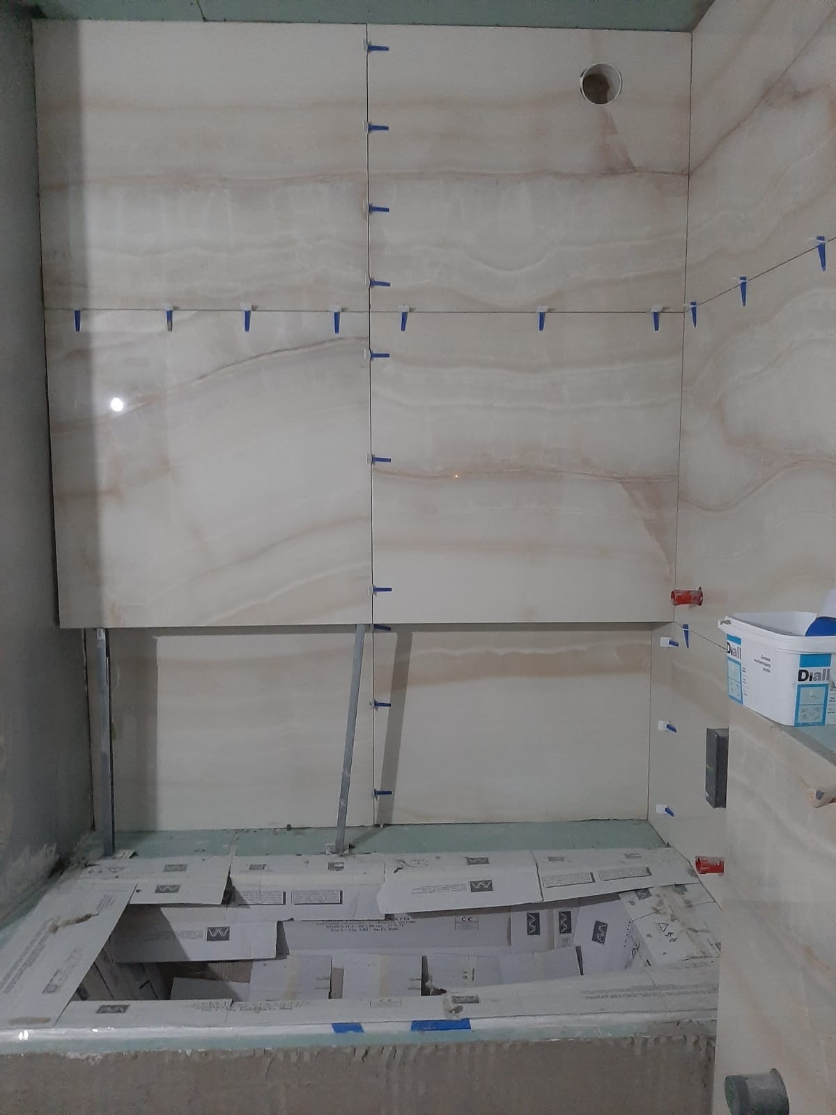 Ściana łazienki w trakcie układania płytek imitujących marmur, z użyciem niebieskich krzyżyków dystansowych i poziomicy, widoczny otwór wentylacyjny w górnej części ściany oraz kartony po płytkach...