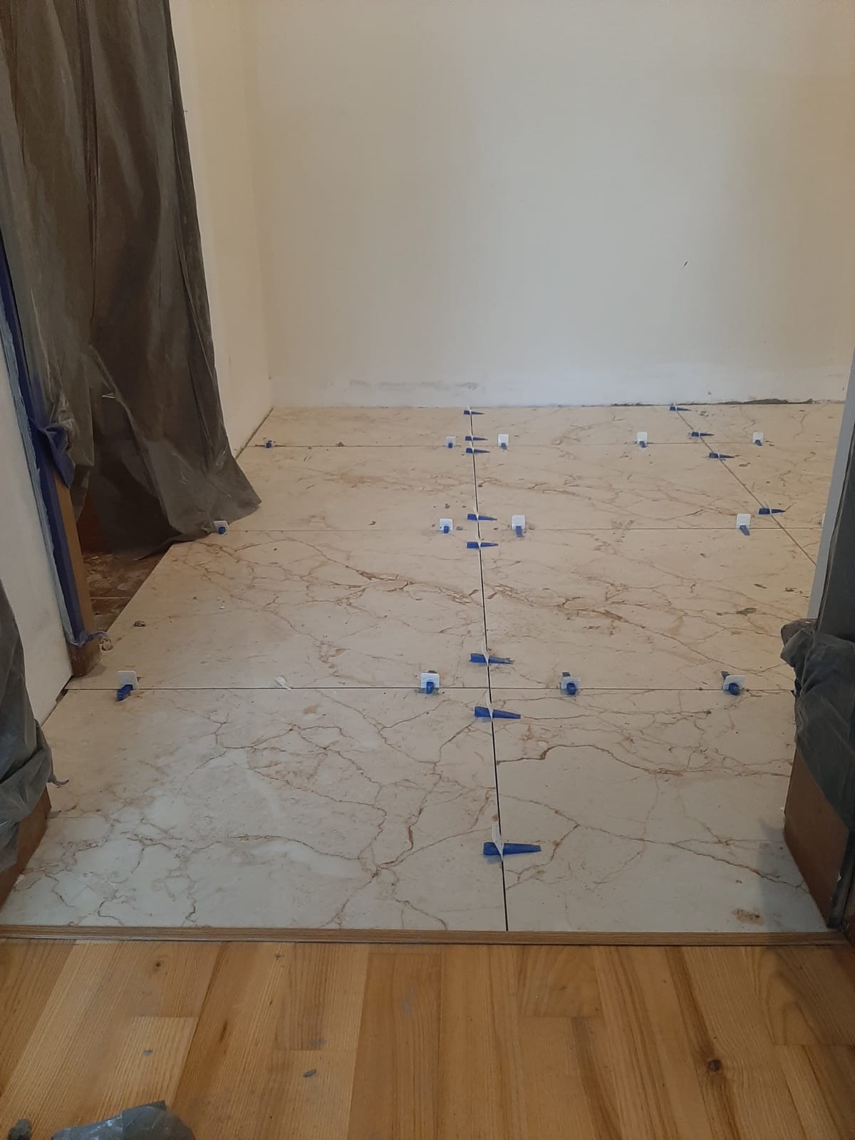 Podłoga z płytek imitujących marmur w trakcie układania, z użyciem niebieskich klinów poziomujących, widok na fragment pomieszczenia z drewnianym progiem.