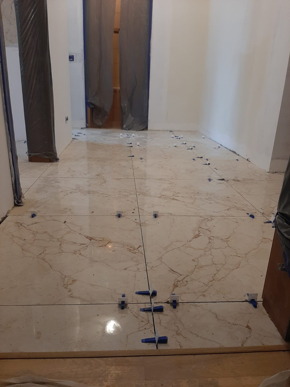 Układanie płytek podłogowych imitujących marmur, z użyciem niebieskich krzyżyków dystansowych, widoczne zabezpieczone folią drzwi w tle.