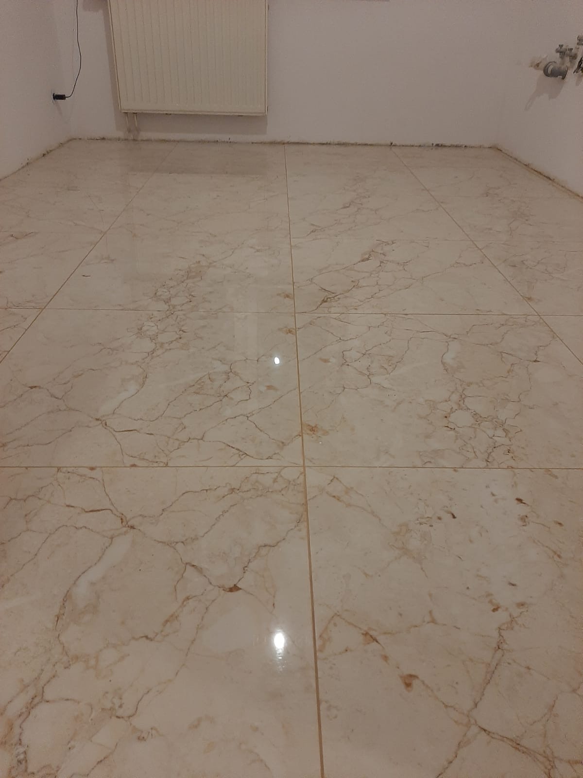 Wykończona podłoga z płytek imitujących marmur w jasnych odcieniach beżu i brązu, widoczne fugi i fragment grzejnika na ścianie.