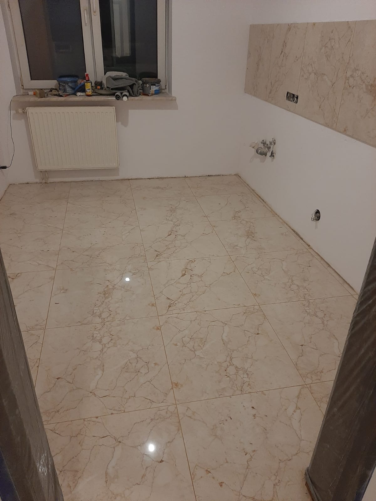 Wykończona podłoga z beżowych płytek imitujących marmur w łazience w Warszawie, widoczne narzędzia i materiały budowlane na parapecie okna.