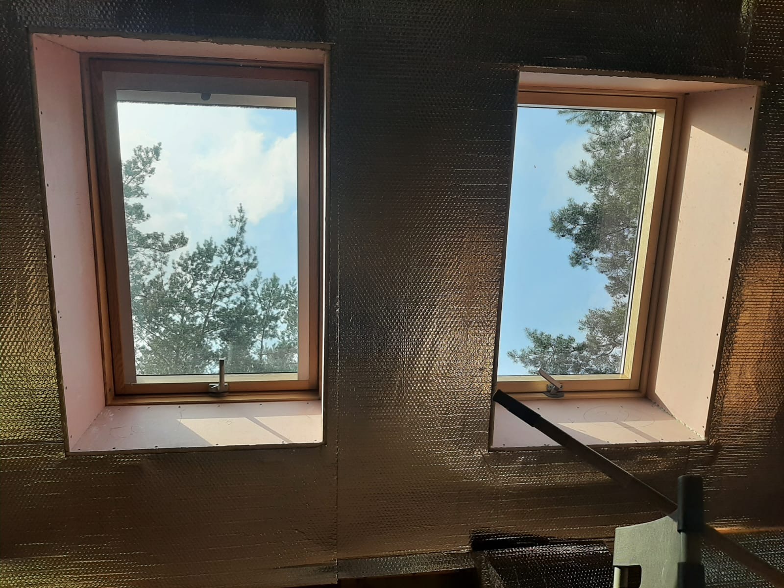 Dwa okna dachowe z widokiem na drzewa i błękitne niebo, ściany pokryte folią termoizolacyjną, fragment drewnianej konstrukcji w dolnej części kadru.