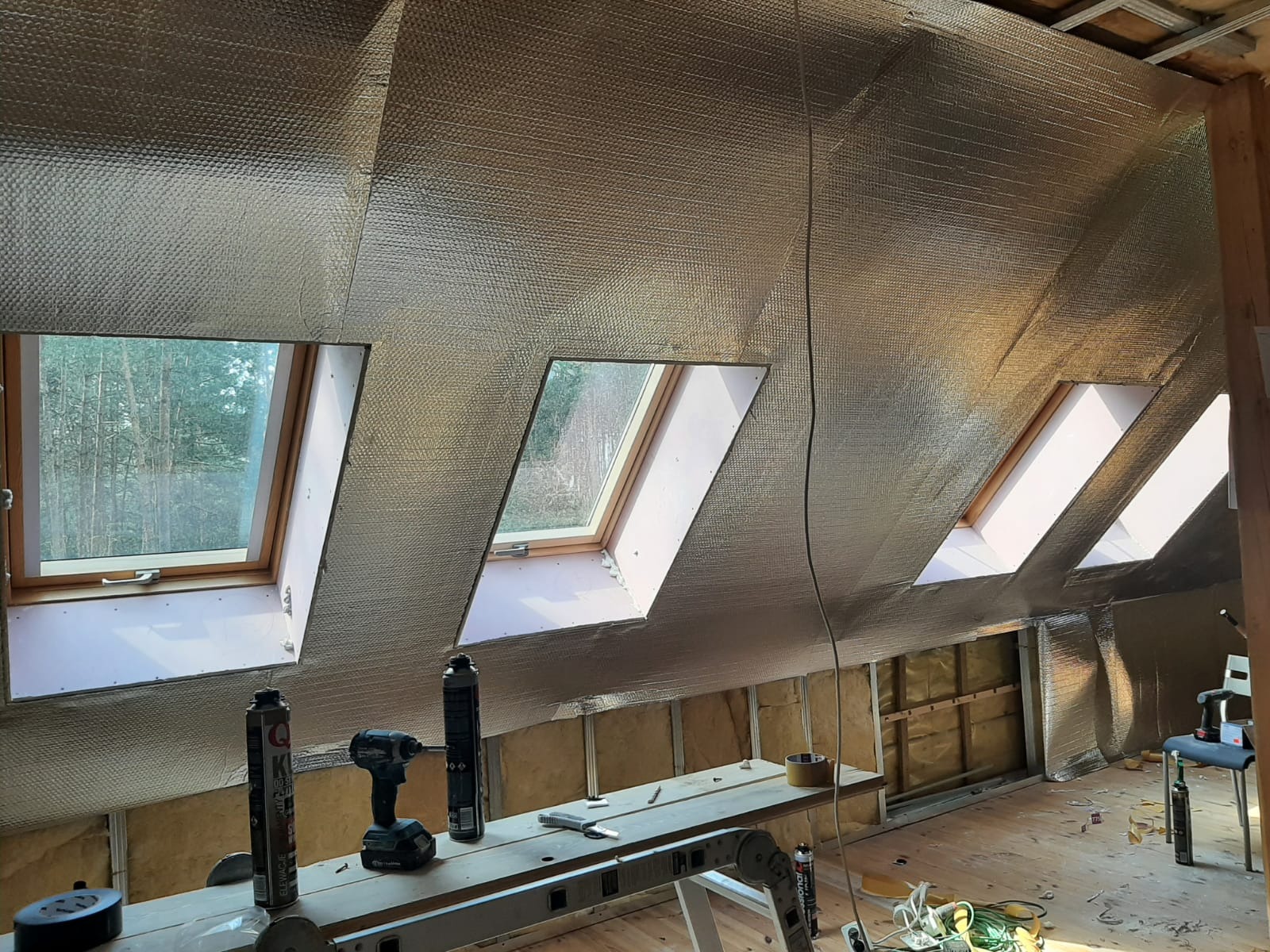 Izolacja poddasza folią termoizolacyjną z widocznymi oknami dachowymi, narzędzia na drabinie, widok na las za oknami.