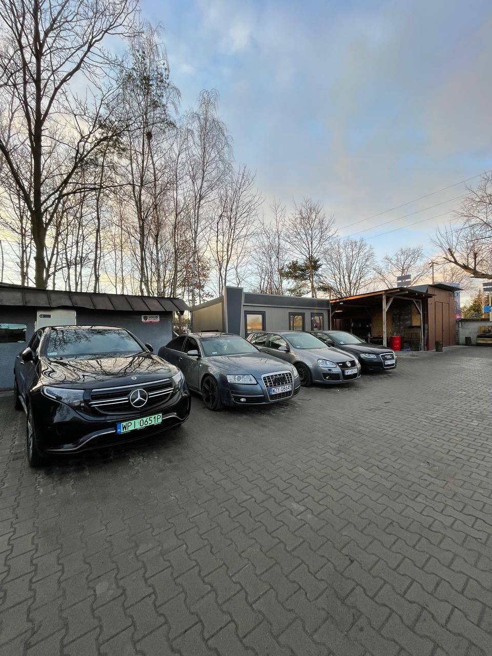 Rząd zaparkowanych samochodów różnych marek, w tym czarny Mercedes EQB z widoczną tablicą rejestracyjną, przed budynkiem warsztatu samochodowego z szarym kontenerem biurowym i zadaszoną wiatą.