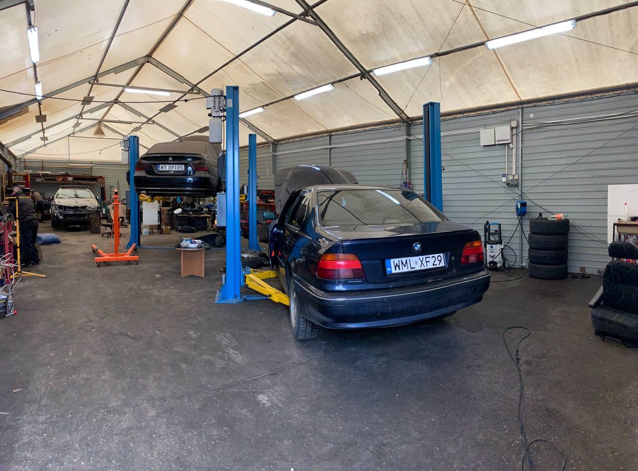 Wnętrze warsztatu samochodowego z podnośnikami, na jednym z nich uniesiony czarny samochód, a na drugim ciemny sedan BMW z otwartą maską, widoczne opony i narzędzia.