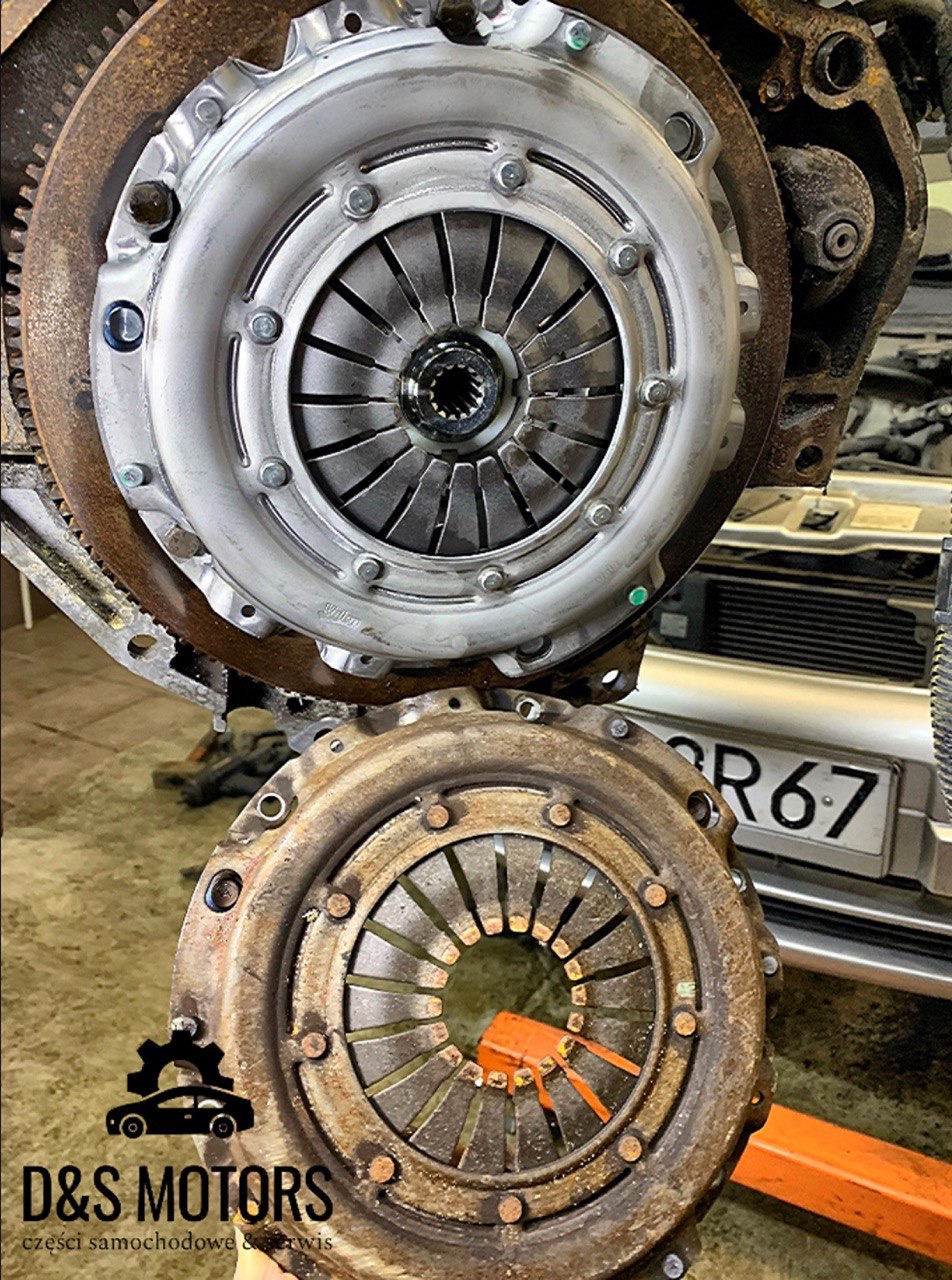 Nowe sprzęgło zamontowane obok starego, zużytego sprzęgła samochodowego na tle fragmentu samochodu z widoczną tablicą rejestracyjną.