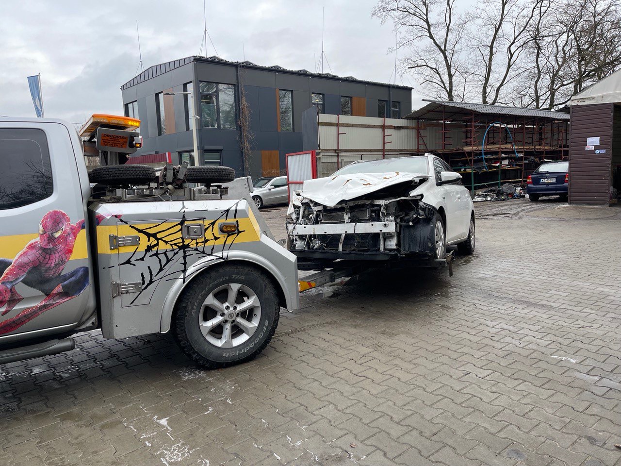 Uszkodzony biały samochód na lawecie holowniczej z grafiką Spider-Mana i motywem pajęczyny, zaparkowany na brukowanym placu przed warsztatem.