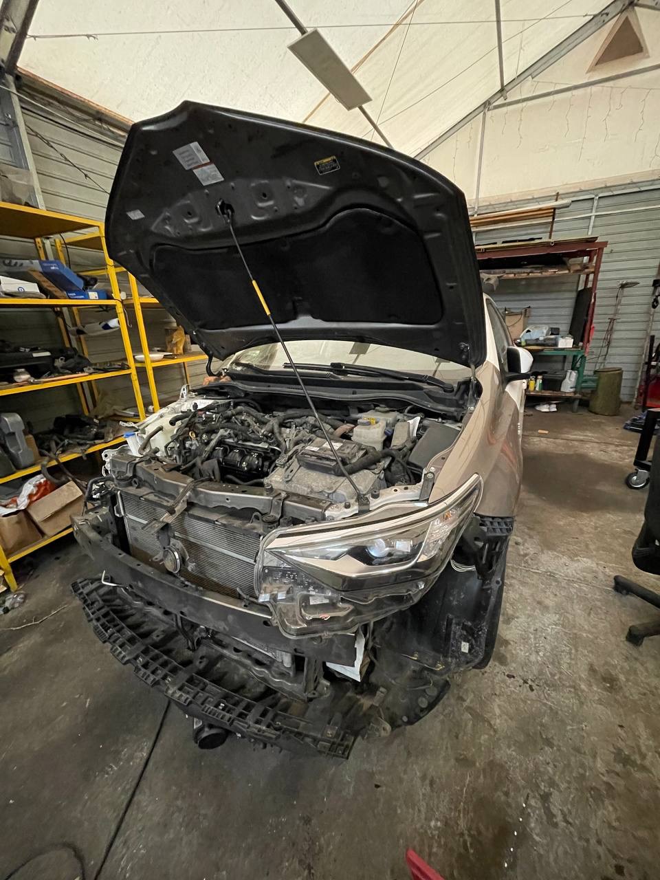 Brązowy samochód z otwartą maską w warsztacie, widoczne elementy silnika i zdemontowany przedni zderzak.