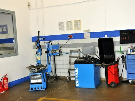 Wnętrze warsztatu samochodowego z niebieskimi urządzeniami do wymiany opon i wyważania kół, widoczne narzędzia i akcesoria, ściany w biało-niebieskiej kolorystyce.