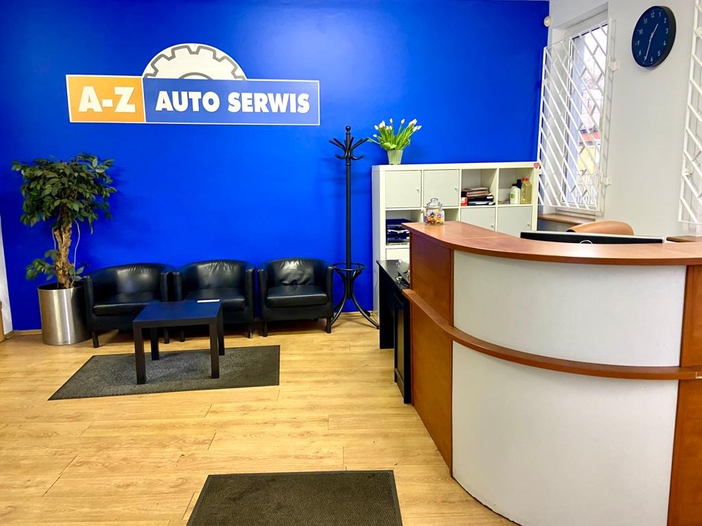 Poczekalnia w auto serwisie: niebieska ściana z logo A-Z Auto Serwis, trzy czarne fotele klubowe, stolik kawowy, recepcja z drewnianym blatem, roślina doniczkowa i bukiet tulipanów na szafce.