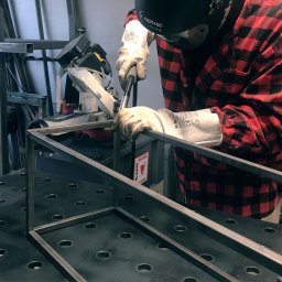 Woofie - Pracownik w czerwonej koszuli w kratę spawa metalową konstrukcję na perforowanym stole spawalniczym, używając maski spawalniczej Optrel.