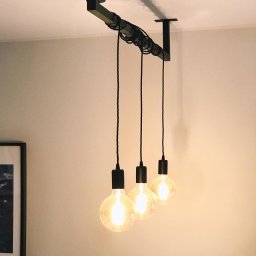 Woofie - Minimalistyczna lampa sufitowa z trzema żarówkami Edisona zawieszonymi na czarnych kablach, przymocowanych do metalowej belki, oświetlona ciepłym światłem, widoczna na tle jasnej ściany.
