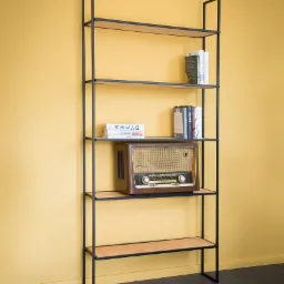 Wysoki, nowoczesny regał z czarną metalową ramą i jasnymi drewnianymi półkami, na których stoją książki i zabytkowe radio, na tle żółtej ściany.