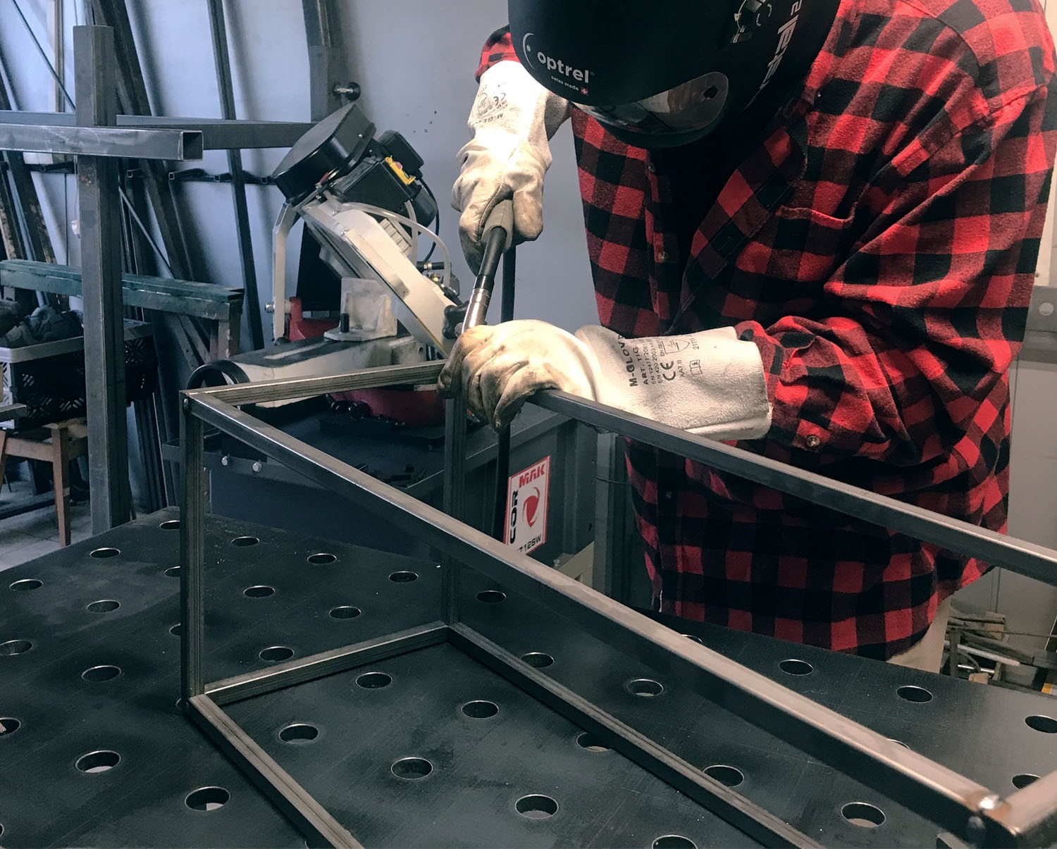 Pracownik w czerwonej koszuli w kratę spawa metalową konstrukcję na perforowanym stole spawalniczym, używając maski spawalniczej Optrel.